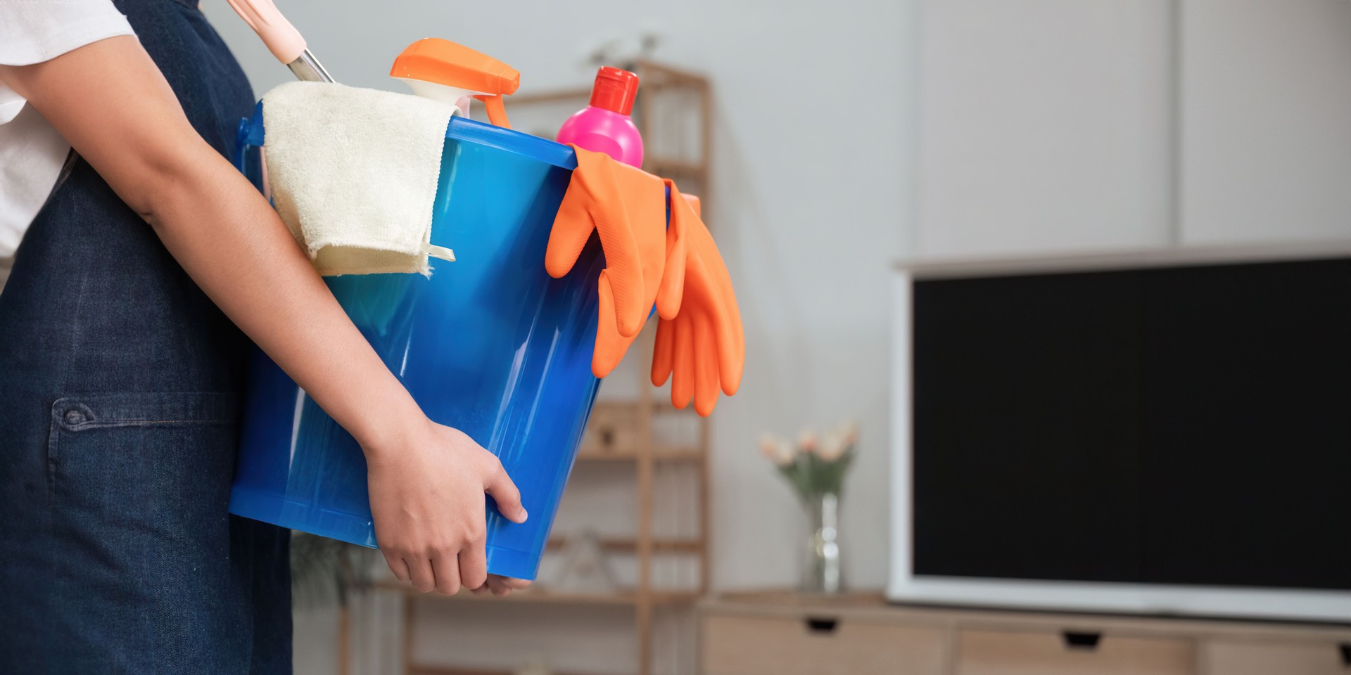 A cleaning woman is standing inside a building holding a blue bucket fulfilled with chemicals and facilities for tidying up in her hand.